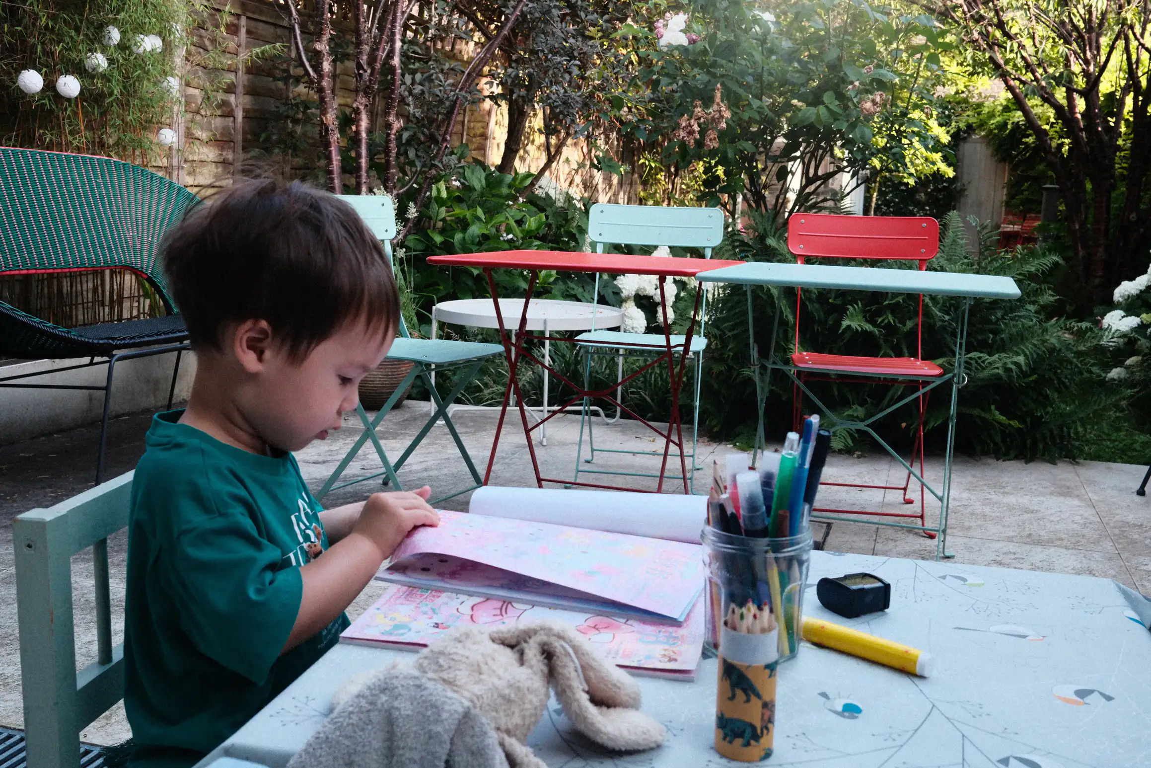 A boy sits at a small table, in front of him a colouring book and a pot of pens, a garden table and chairs can be seen in the background