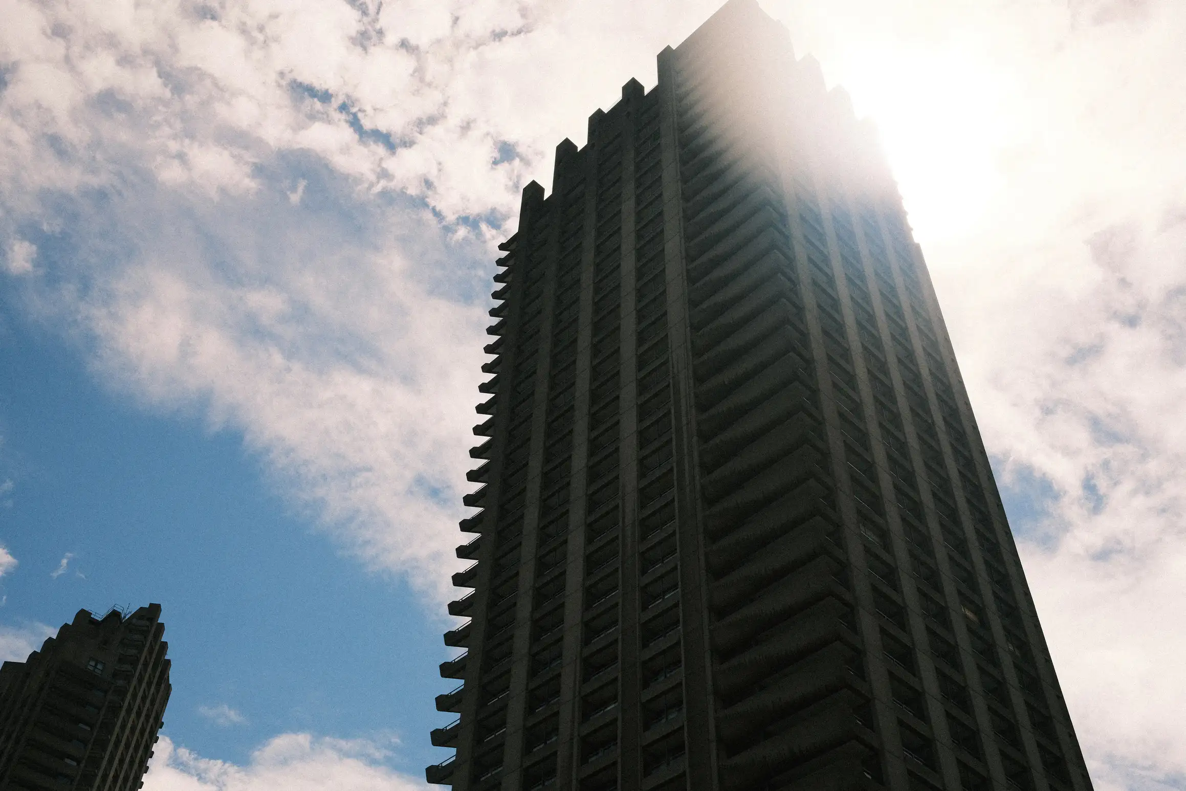 A photo looking up at the midday sky. A tall skyscraper in the foreground. The bright sunlight blooms over the edge of the building. 