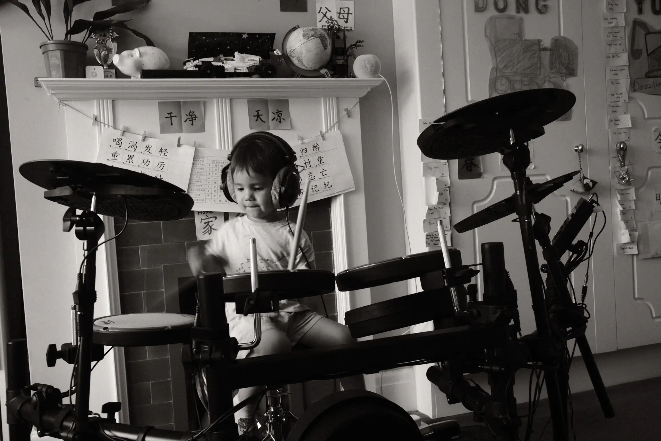 A black and white photo of a young boy playing an electronic drum kit. On the wall behind him, Chinese characters hang from a piece of string.