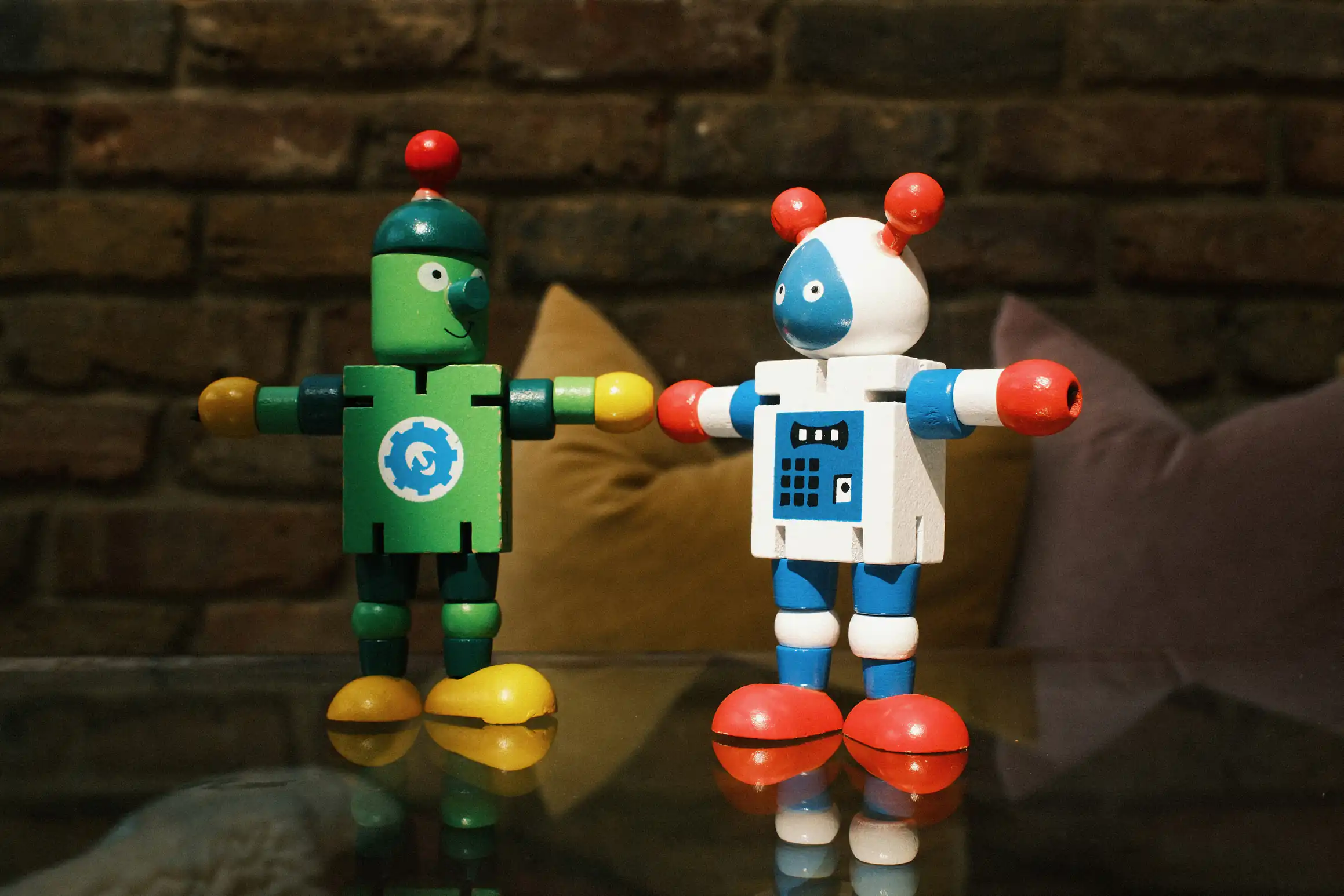 A colour photo of two wooden robotic figures standing on a glass table. Their arms are positioned as if reaching out to each other.