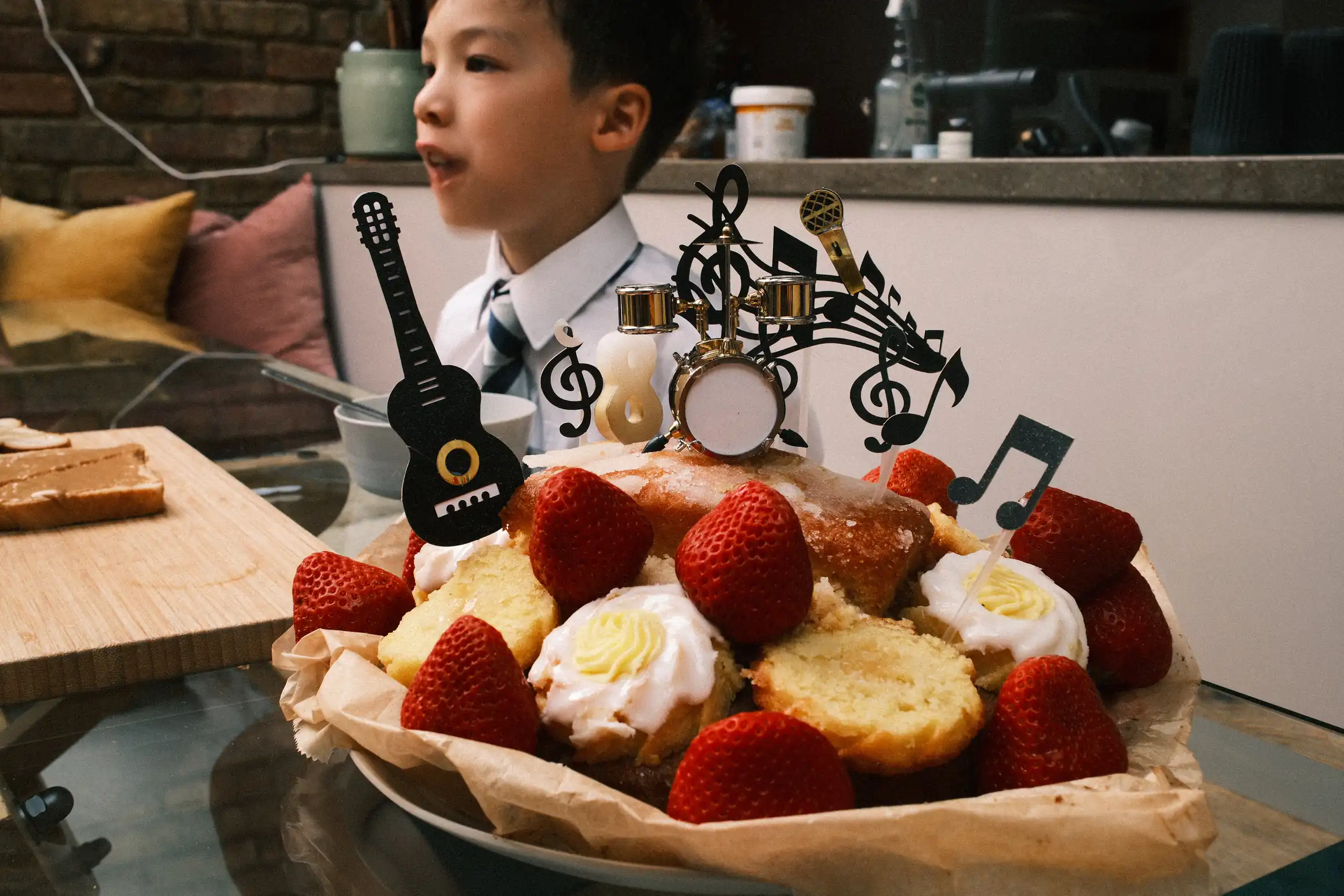 A photo of a birthday cake. The cake is sponge with lemon icing. It is decorated with strawberries, musical notes, a guitar and a drum kit. The candle indicates it is an eighth birtday cake. Behind it a boy looks out of the frame. We don't see what he is looking at.