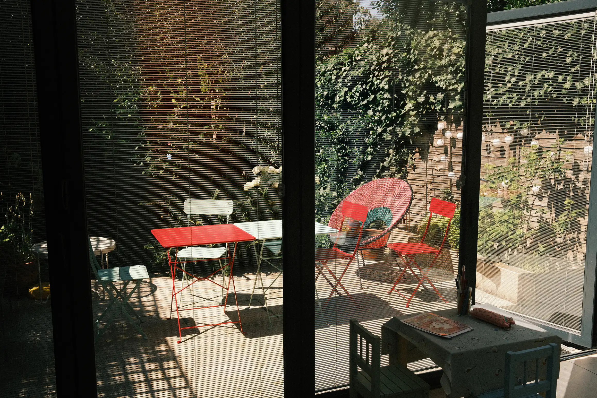 A photo looking out through glass doors to a garden. Despite the blinds being closed, the bright sun outside lights the garden leaving the room in the dark.