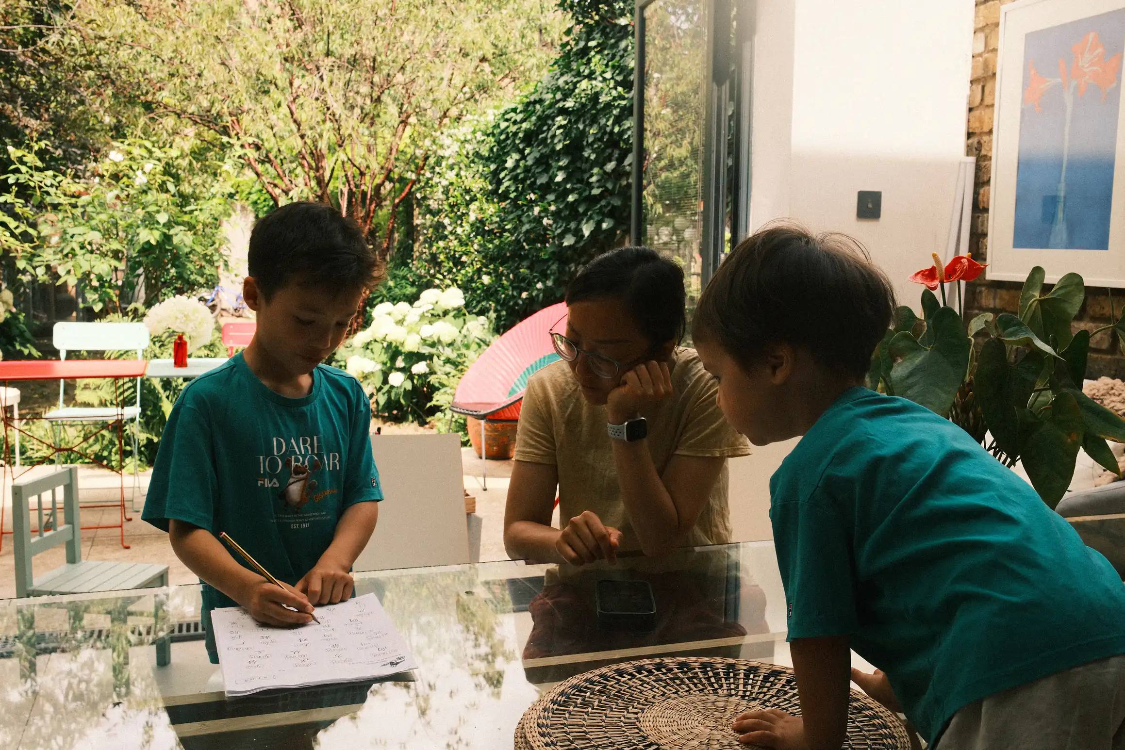 A young boy stands at a table with a pencil in hand, writing on a piece of paper. A woman looks on, head resting on her hand. In the foreground a younger boy leans over the table to see what is being written.