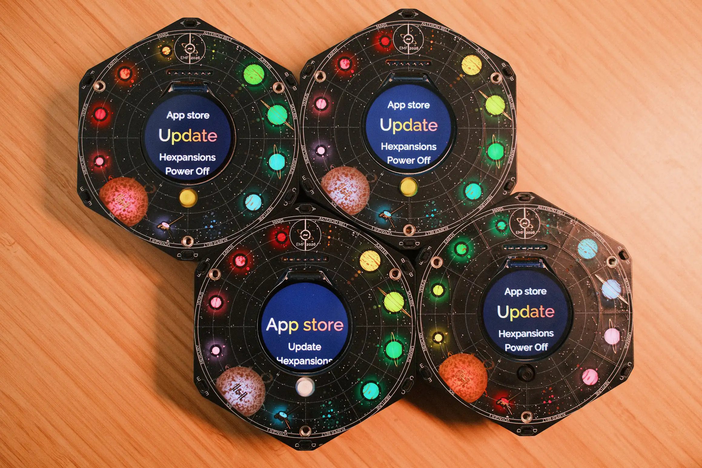 A photo of four EMF Camp Badges. The badges are hexagons, arranged with one side touching each other. The badges are all powered on and showing the main menu with 'Update' selected.