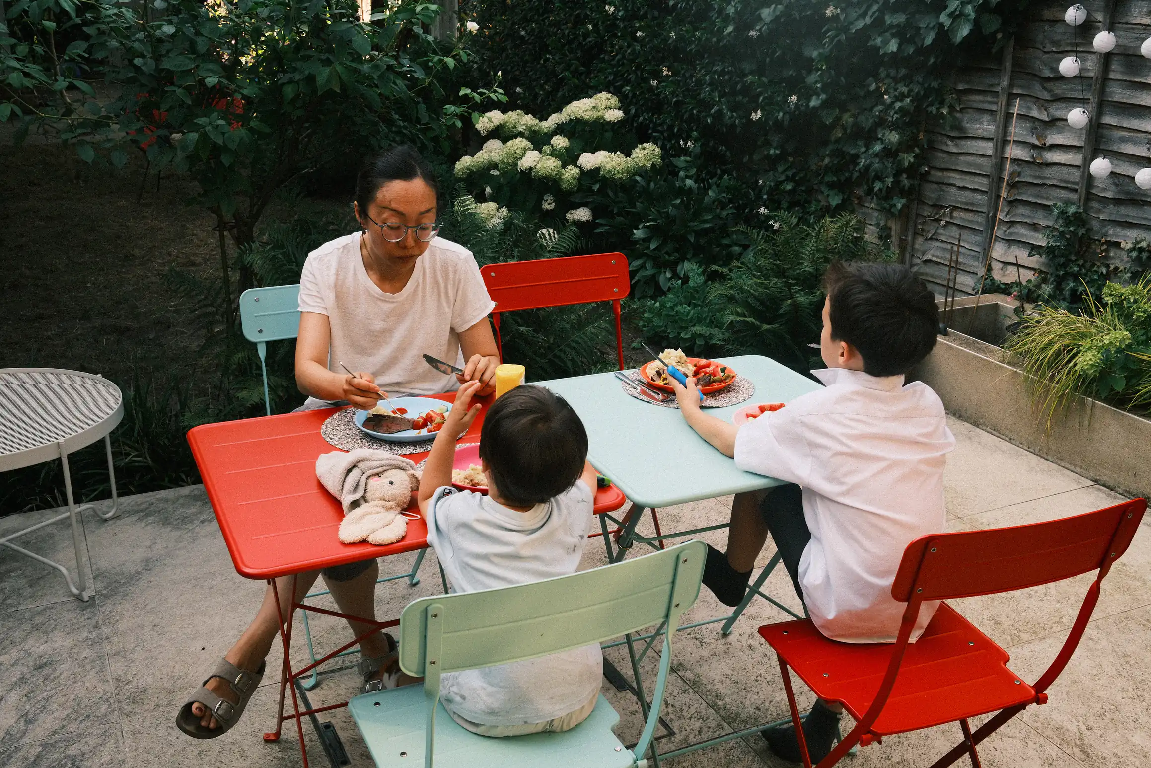 Three people sit at two tables in a garden. There is food on the table in front of them. A fourth place is laid out but no one sits there.