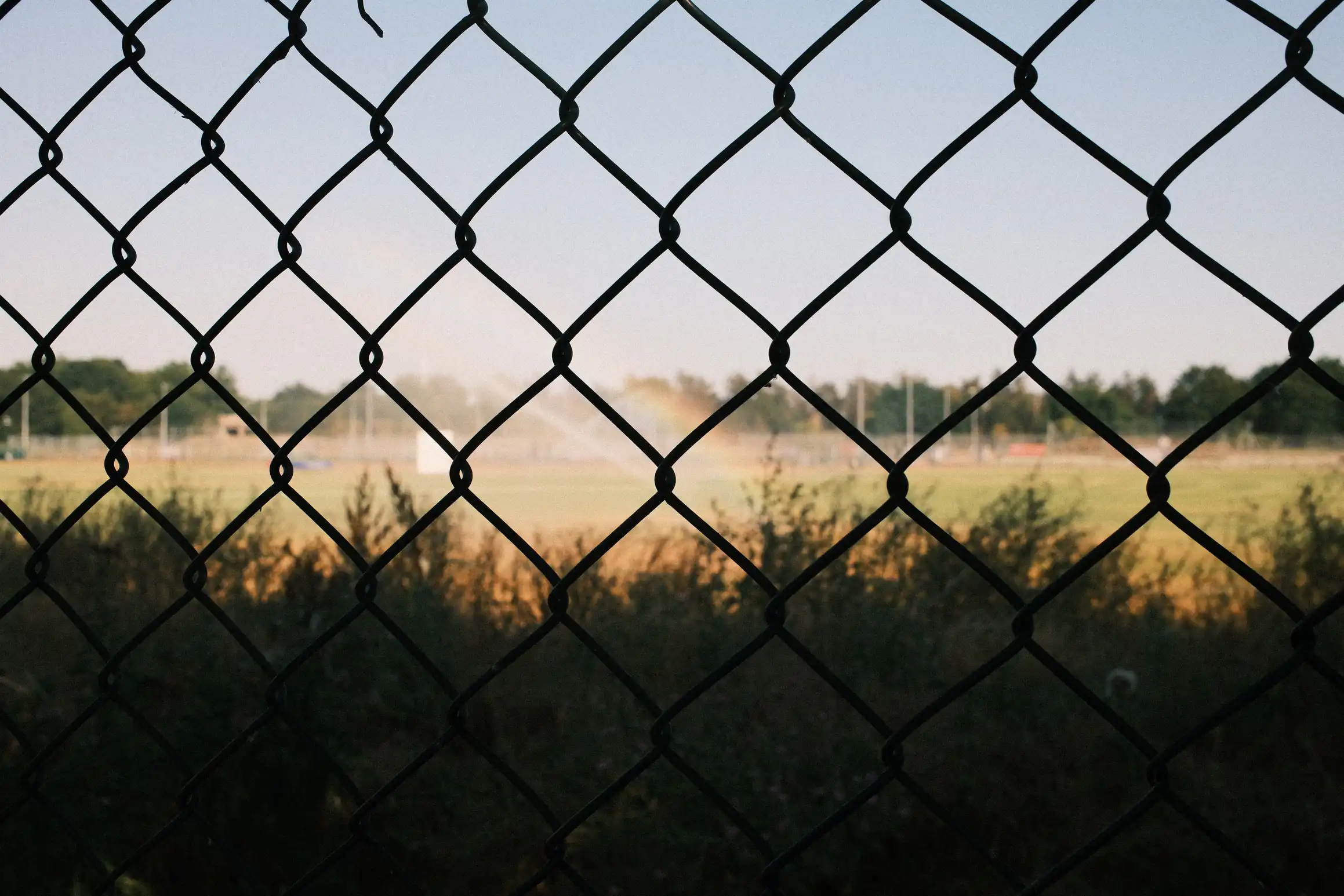 A photo taken through some wire fence. Behind the fence, in the shade, some wild grass. In the distance, brightly lit playing fields. A subtle rainbow appears in the spray of a sprinkler.