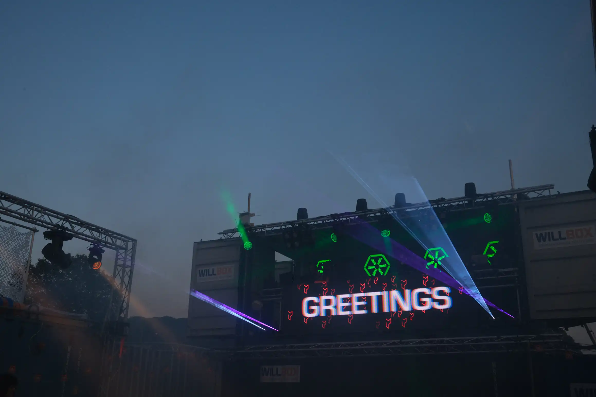 A nighttime photo looking up at a DJ booth above a dance floor. Lights shine towards the photographer. A screen on the front of the booth has the word greetings in white letters.