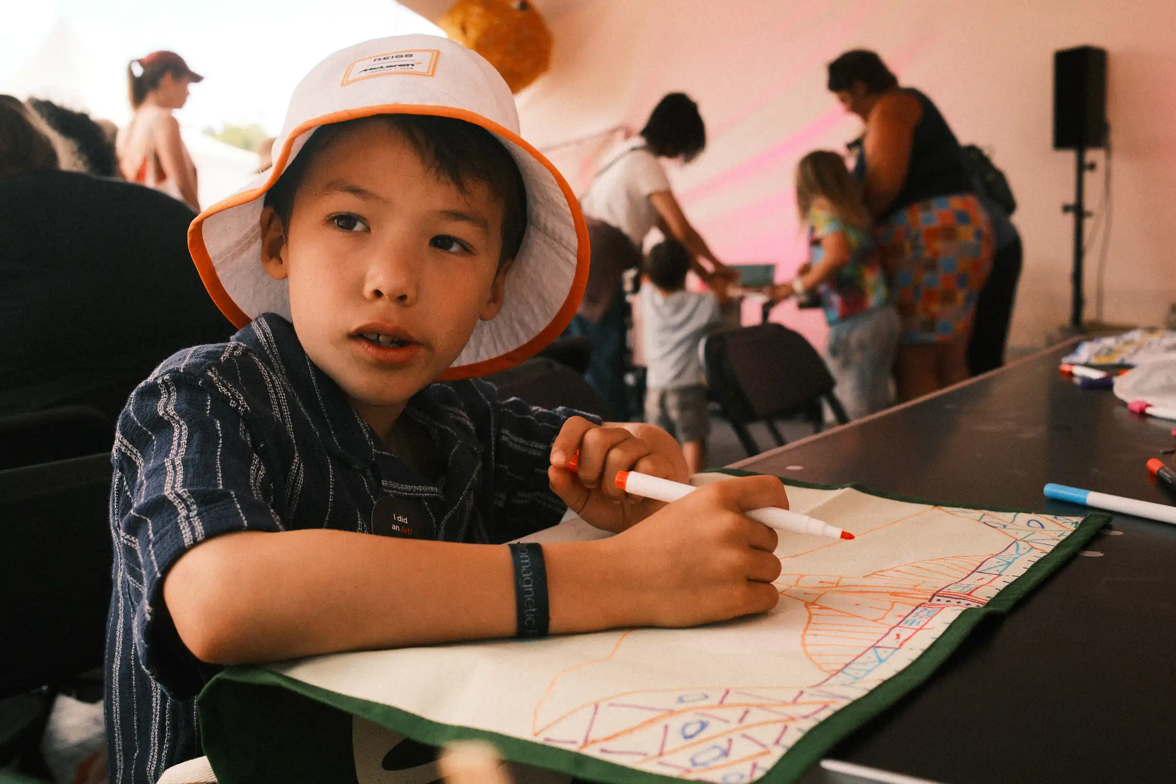 A boy looks off into the distance behind the photographer. In his hand he holds a felt tip pen in front of him on a table. An upturned tea towel. Lines mark out a new design on the back.