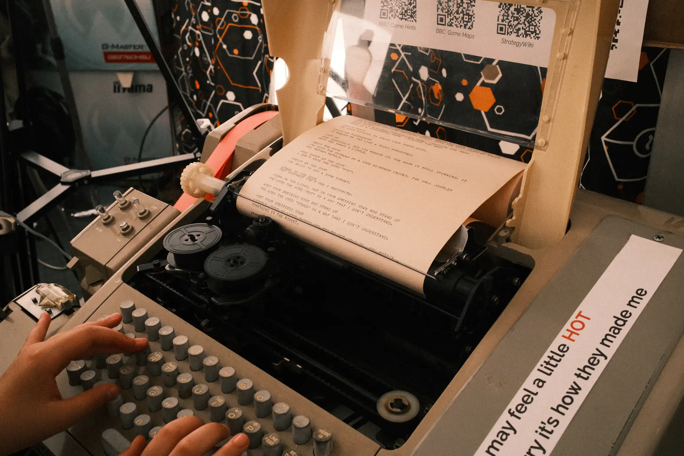A photo of a teletype machine two hands type on the keyboard in front of the machine several QR codes provide links to an explanation of the game on the piece of paper that appears from the machine instructions a text-based adventure game