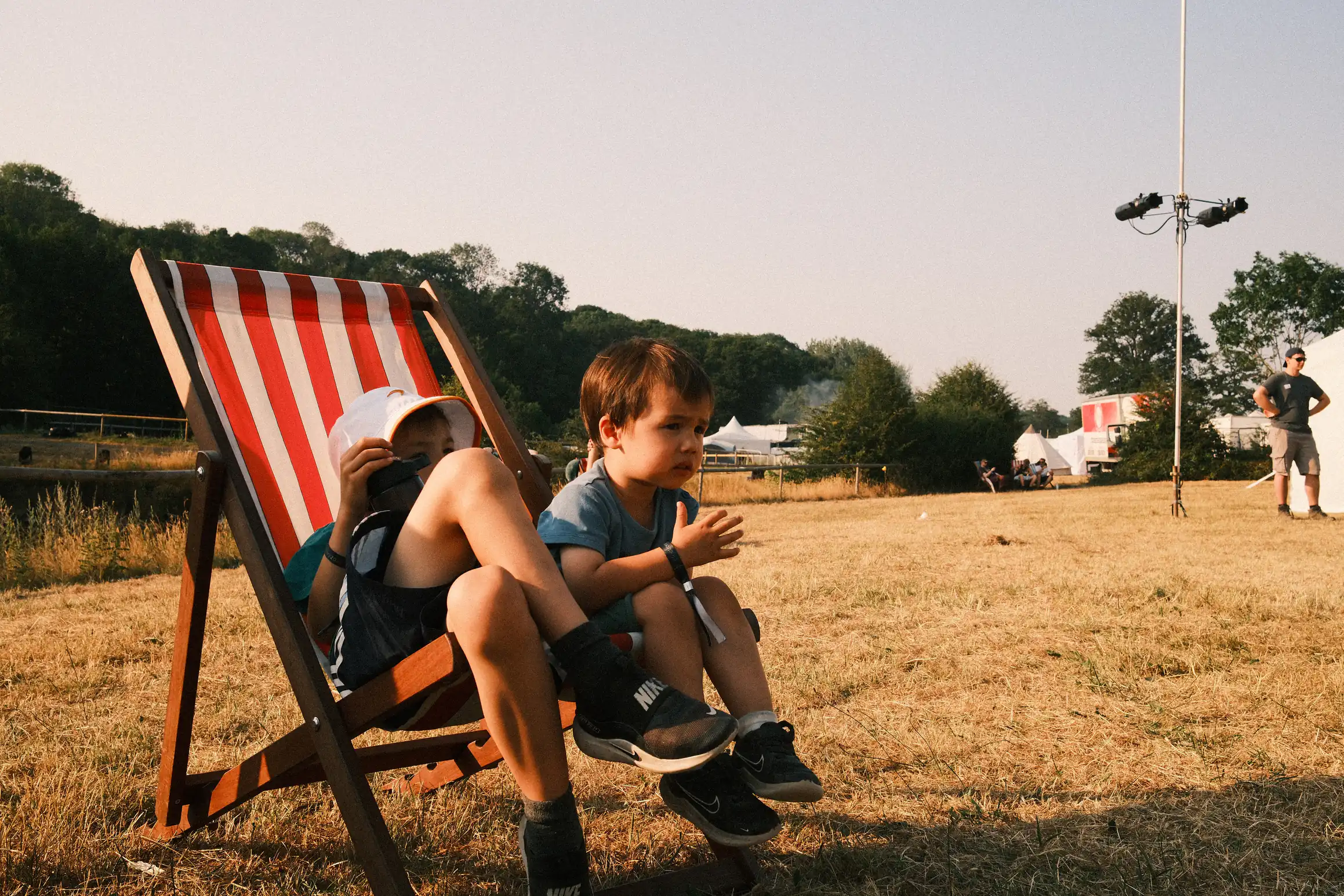 Two boys sit on deck chairs in an otherwise empty looking field. The grass is dry and yellow 