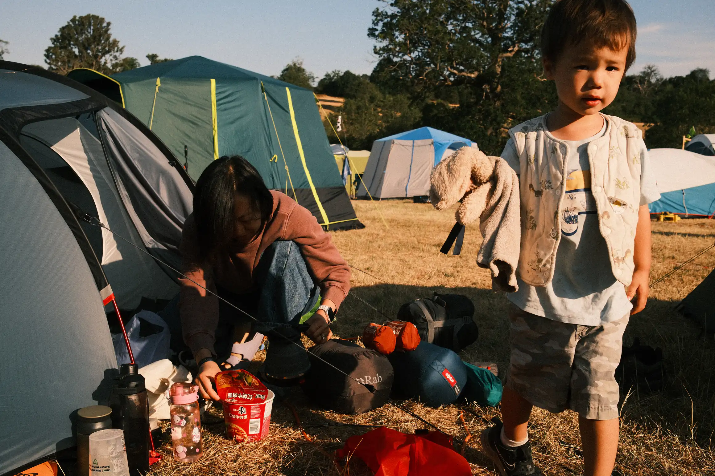 A young boy holds his rabbit walking away from a tent by the door of the tent. A person kneels over a pot of instant noodles. Meal preparation in progress 