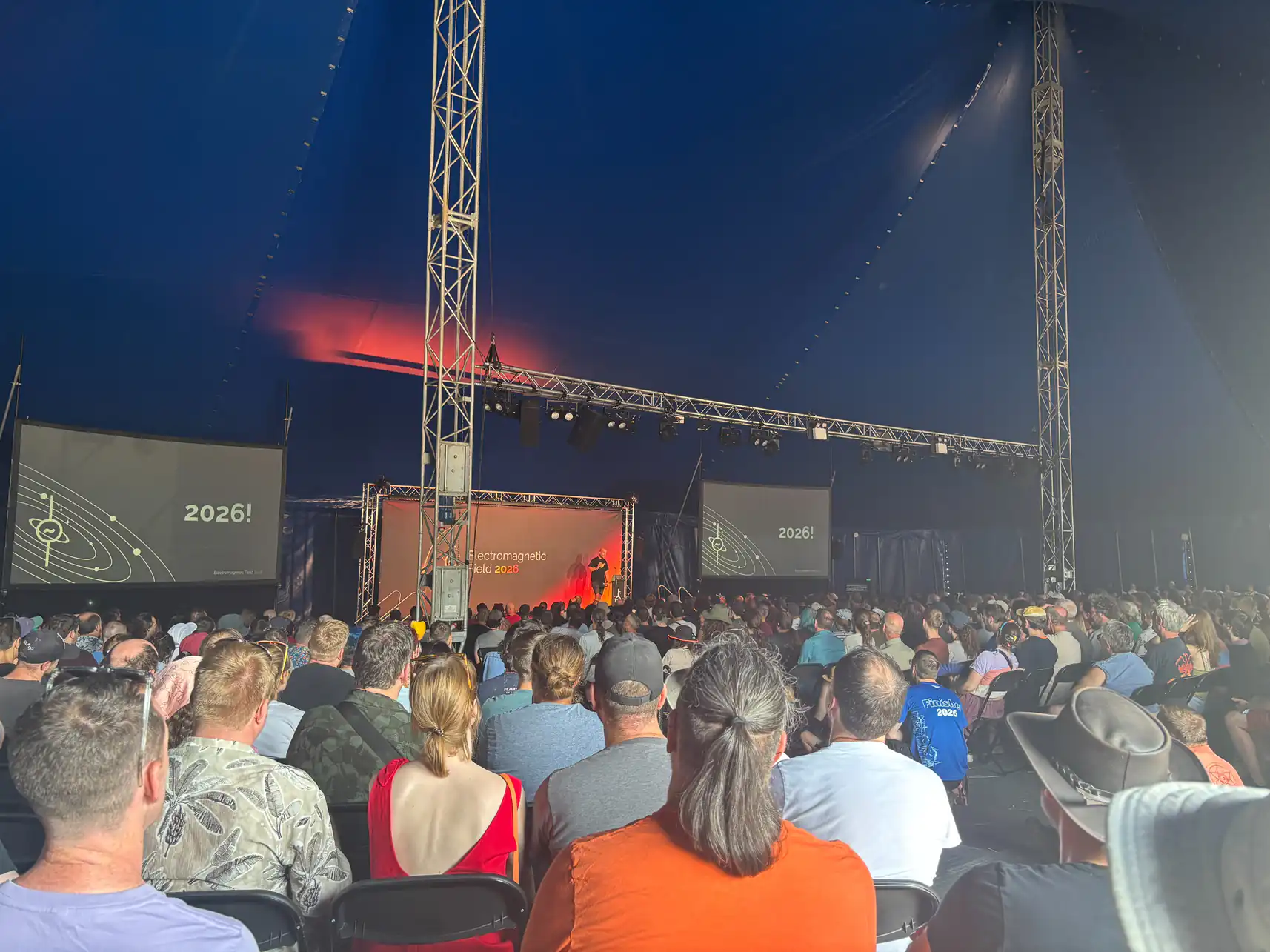 A photo of a marquee tent filled with a crowd of people looking towards a stage. Either side of the stage two large screens. A person stands on stage. Behind them a background, says EMF 