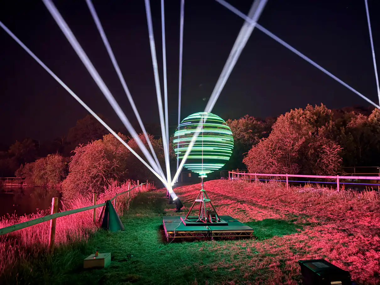 A slow-motion photo captures a spinning globe of LEDs. Behind the globe, searchlights light up the sky. The foreground is lit with a pink glow 