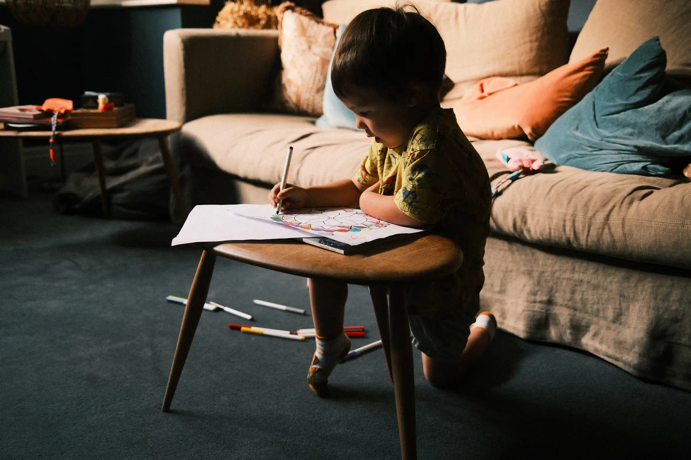 a young boy kneels at a coffee table in front of a sofa, pen in hand, colouring, on the floor behind him, a scattered collection of coloured pens, light from the windows off camera means the majority of the boys figure is silhouted against the bright background