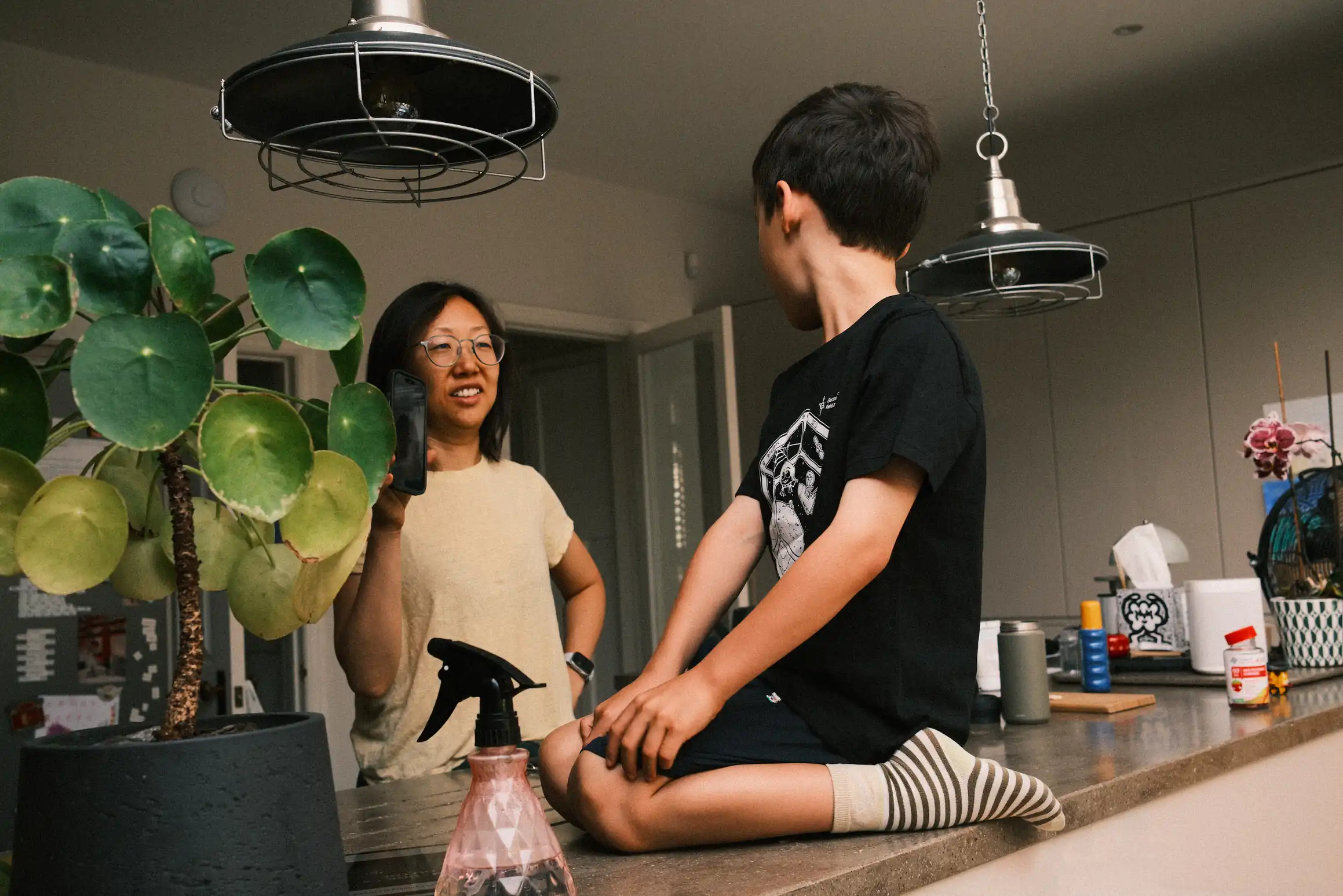 A lady holds up a phone towards a boy, on the screen the image of someone on a video call. The boy is kneeling on a kitchen work surface, in front of him a plant and mister.
