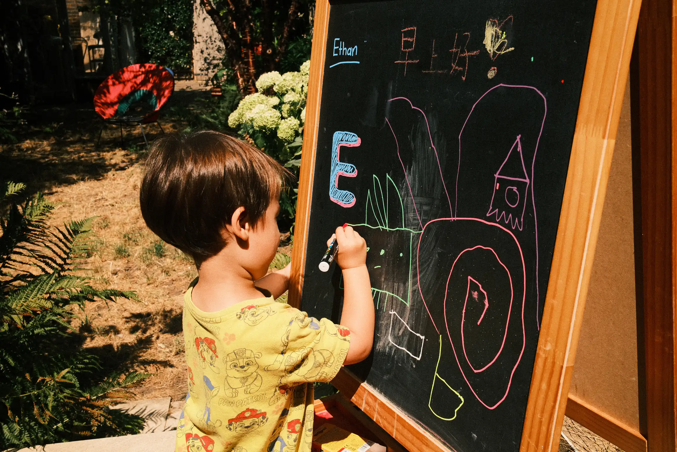 A young boy stands at a chalk board in a garden, he's holding a liquid chalk pen, on the board in front of him are his name, 早上好, and the letter E. He is drawing shapes underneath the writing