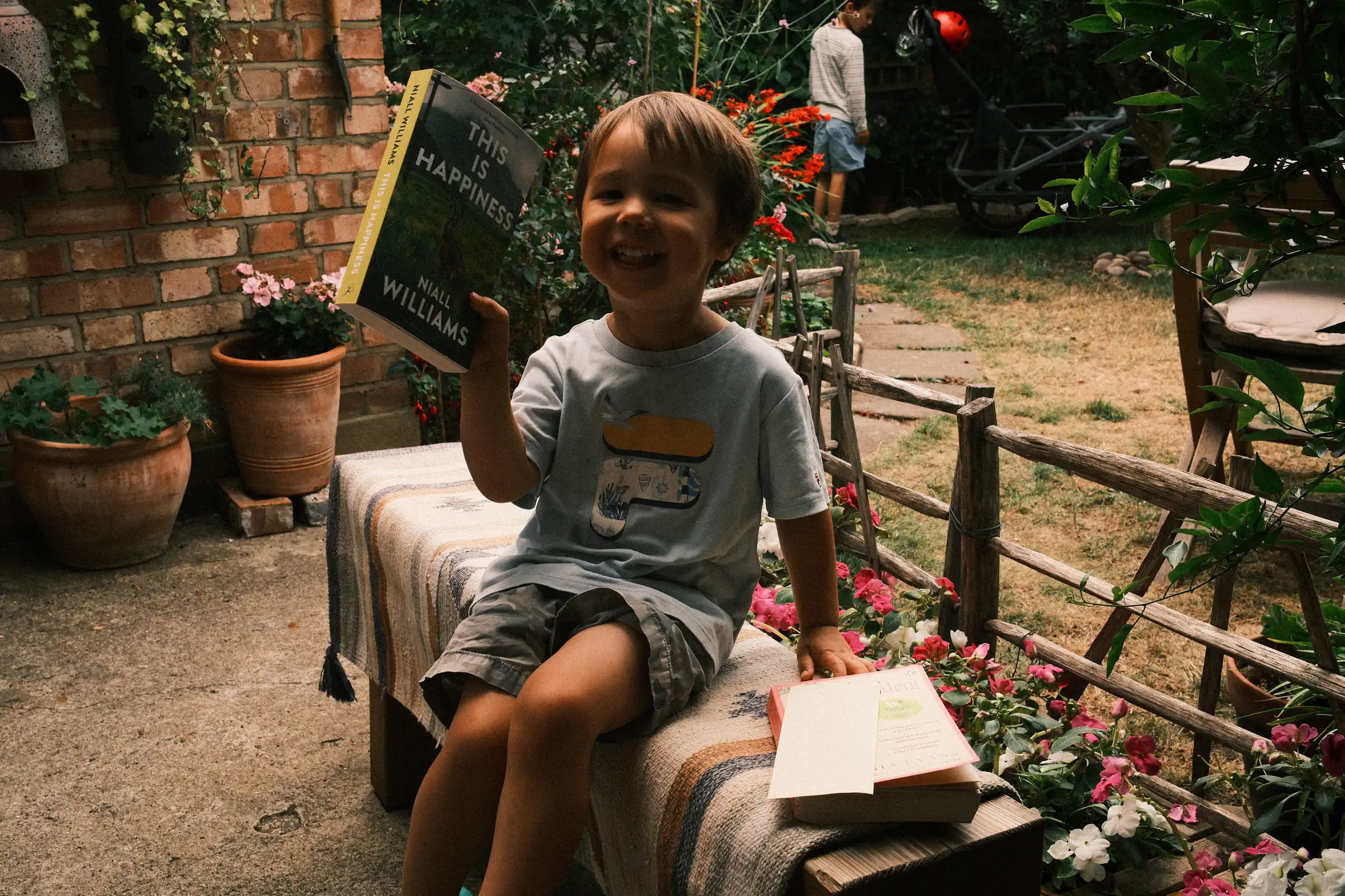 A young boy sits on a garden bench. Behind him, a wooden fence separates the patio area from dry looking grass, in the background another boy stands in the shadows, the boy in the foreground is holding up a book titled, 'this is happiness'.