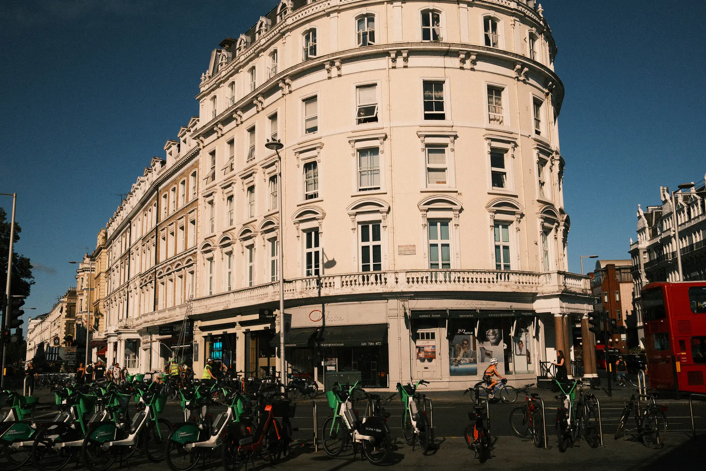 A photo of a white, six story, building outside South Kensington Tube station, the bright sunlight reflecting off the building leaves the foreground in the dark