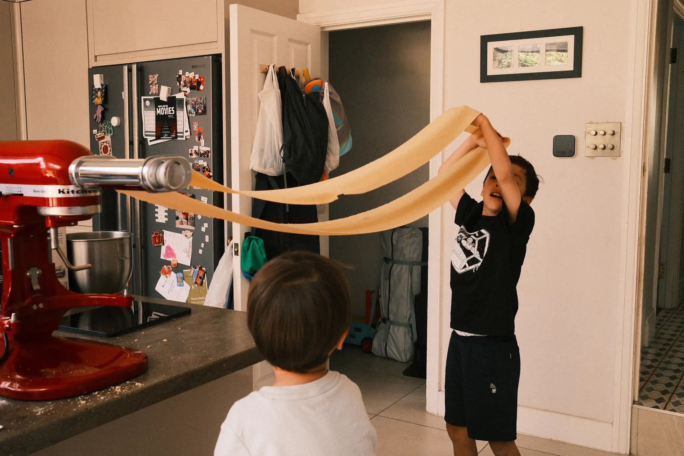 two boys stand in a kitchen making pasta, a long belt of pasta passes through rollers on a pasta machine, the elder opf the two reaches as high as he can to keep the pasta from falling to the floor