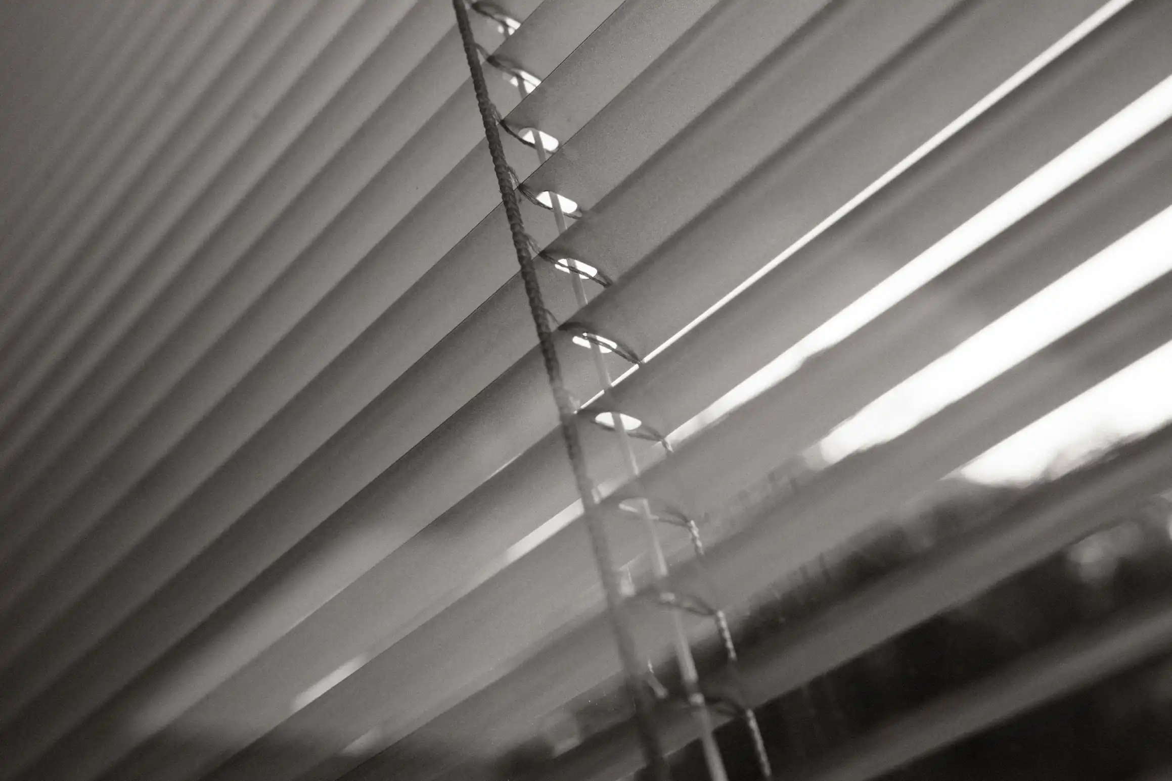 a balck and white photo looking out through the slats of a blind to the bright sky outside.