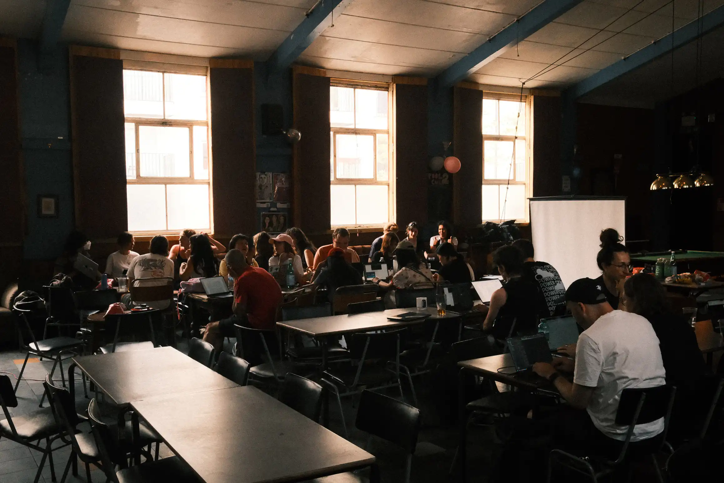 An indoor scene showing tables laid out in a canteen style. People sit at the tables huddled around laptops. The bright lights in the background mean the room appears to be dimly lit.