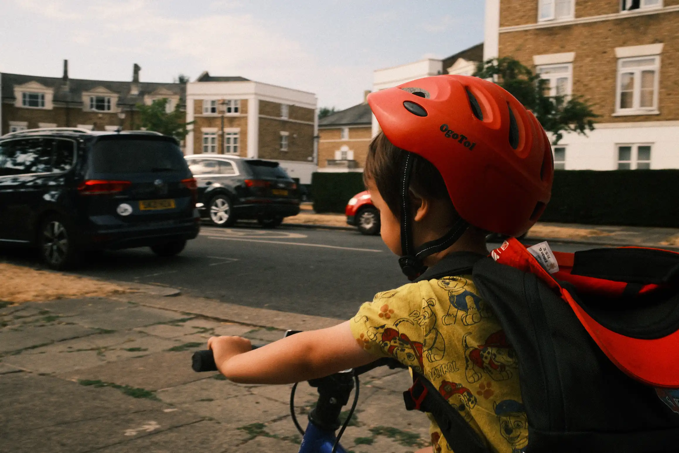 A photo of a young boy cycling along the pavement away from the camera. He is wearing a red helmet. The road behind him is lined with parked cars.