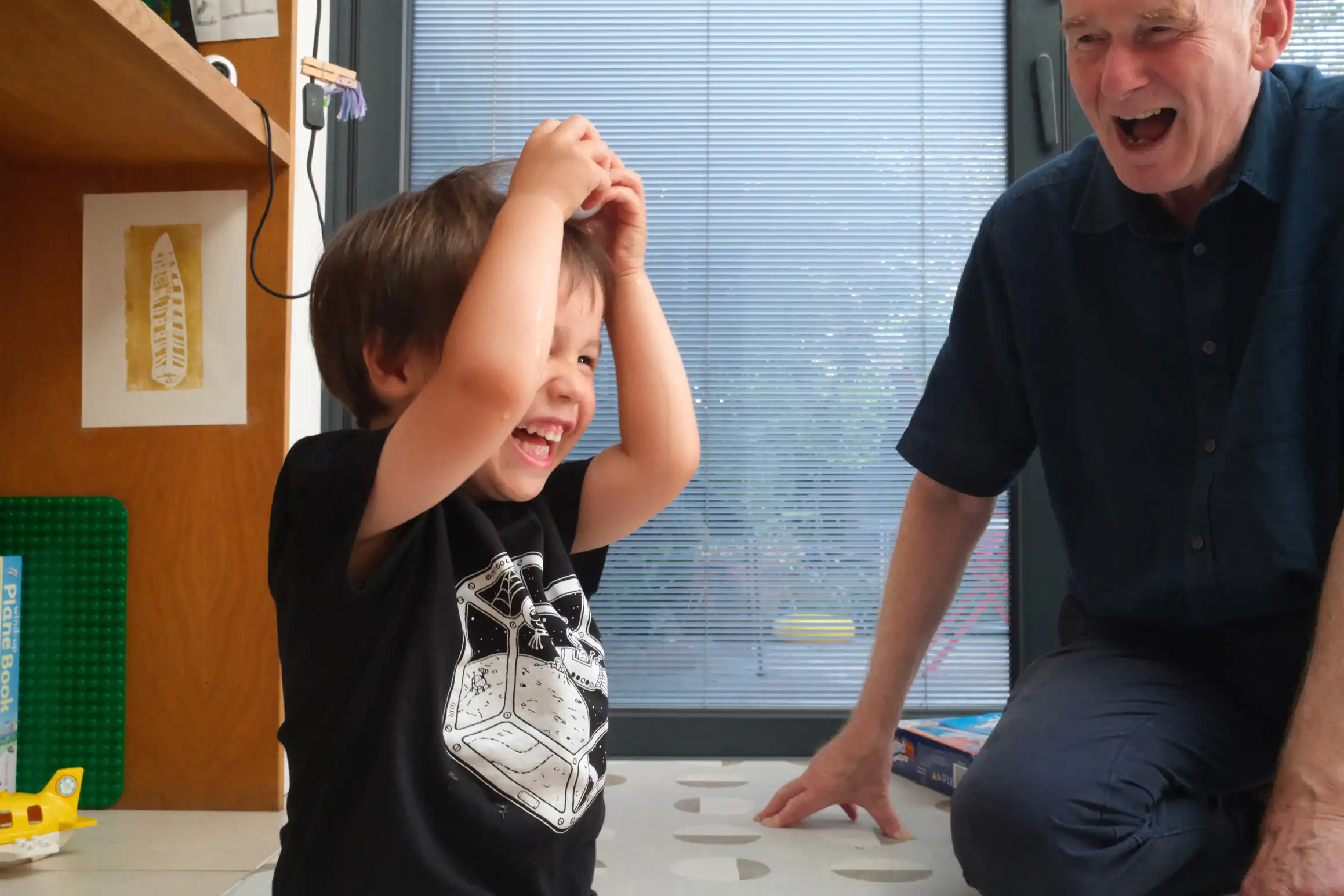 A young boy holds an egg ready to crack it over his head. A man looks on both of them, filled with delight, smiles on their faces.
