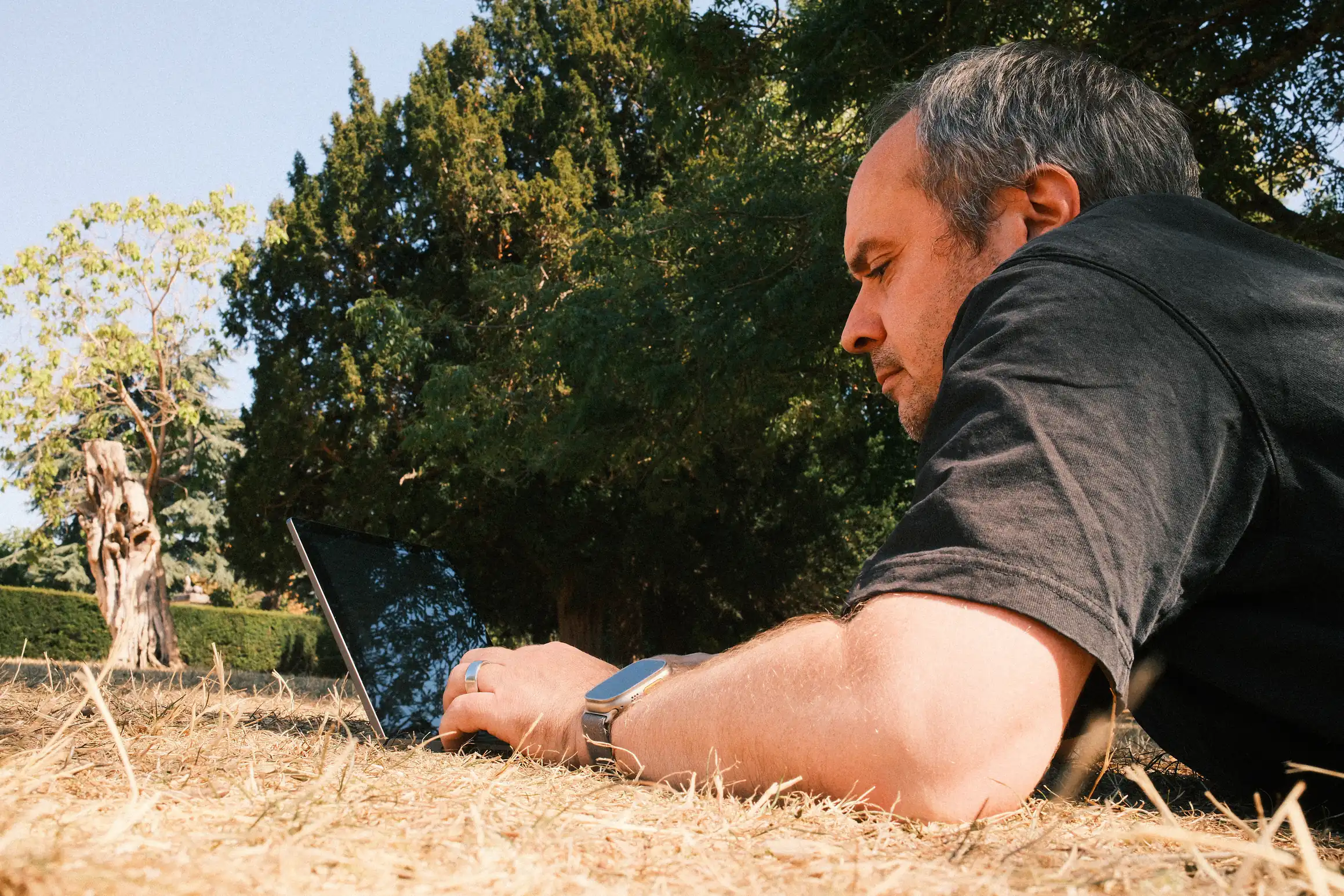 A person lays on dry grass, a laptop in front of him.