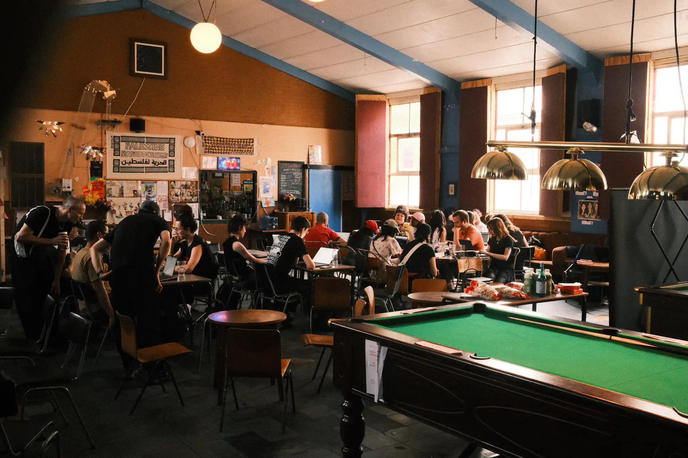 A community centre filled with people they are all sat at long tables looking at laptops. In the foreground a pool table goes unused.