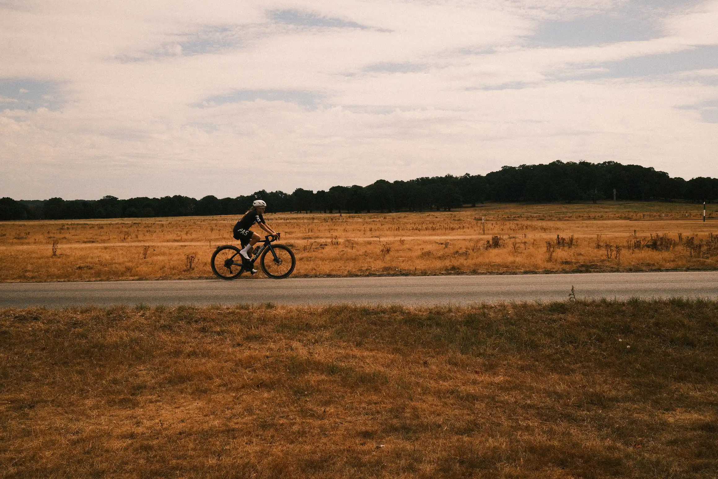 A cyclist rides on an empty road uphill behind them a vast expanse covered in parched yellow grass.