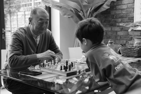 A black and white photo showing two people playing chess. A boy, closest tot he camera, plays black. His grandfather plays white. The grandfather, the least experienced of the two, is smiling as if he has just seen something.