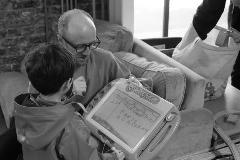 A black and white photo showing two people reviewing a sketch on a children's erasable sketch tablet. A boy faces away from the camera holding the "megasketcher" up to his uncle. The sketch shows a train, with three train cariages. Underneath it, the word "timetable". The first train arives in 22 minutes and is the fast train to Richmond.