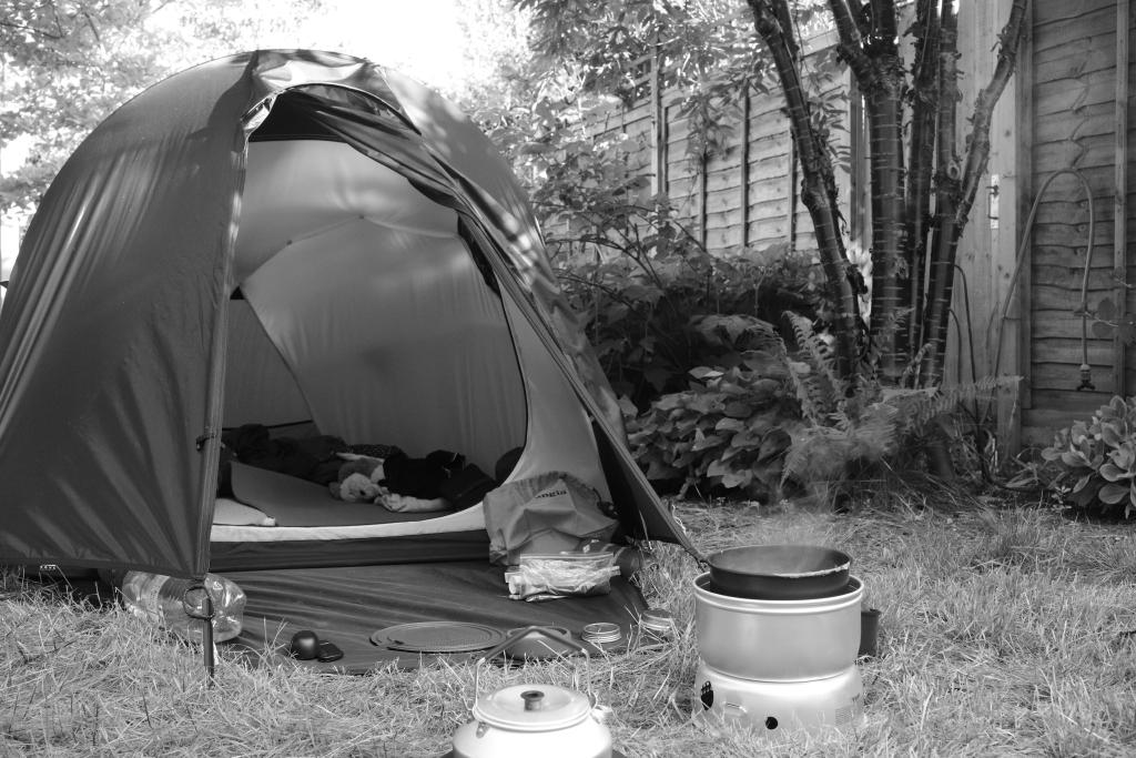 A black and white photo showing a tent in an urban garden. The door to the tent is wide open. In front of the entrance, a cooking stove can be seen with steam coming off the pan.