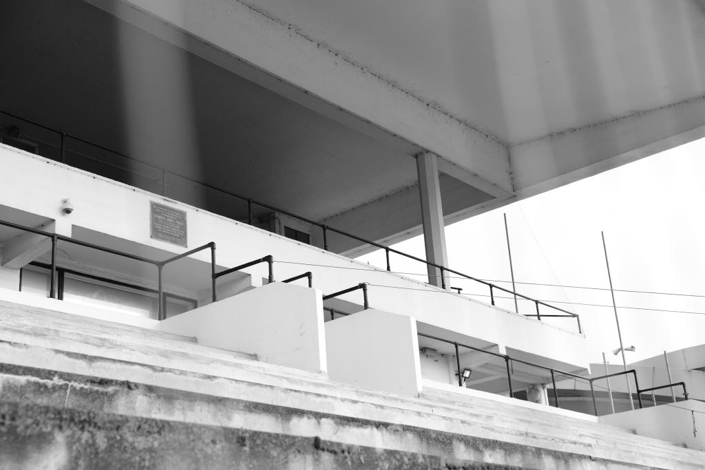 A black and white photo of a concrete sports stadium. The photo looks up towards the roof, the concrete benches visible in the foreground. As you move up the image you see a balcony before moving up towards the second tier. The first tier is over-exposed, washed out by the bright sun. The second tier of seating is much darker, shaded by the concrete roof of the stadium.