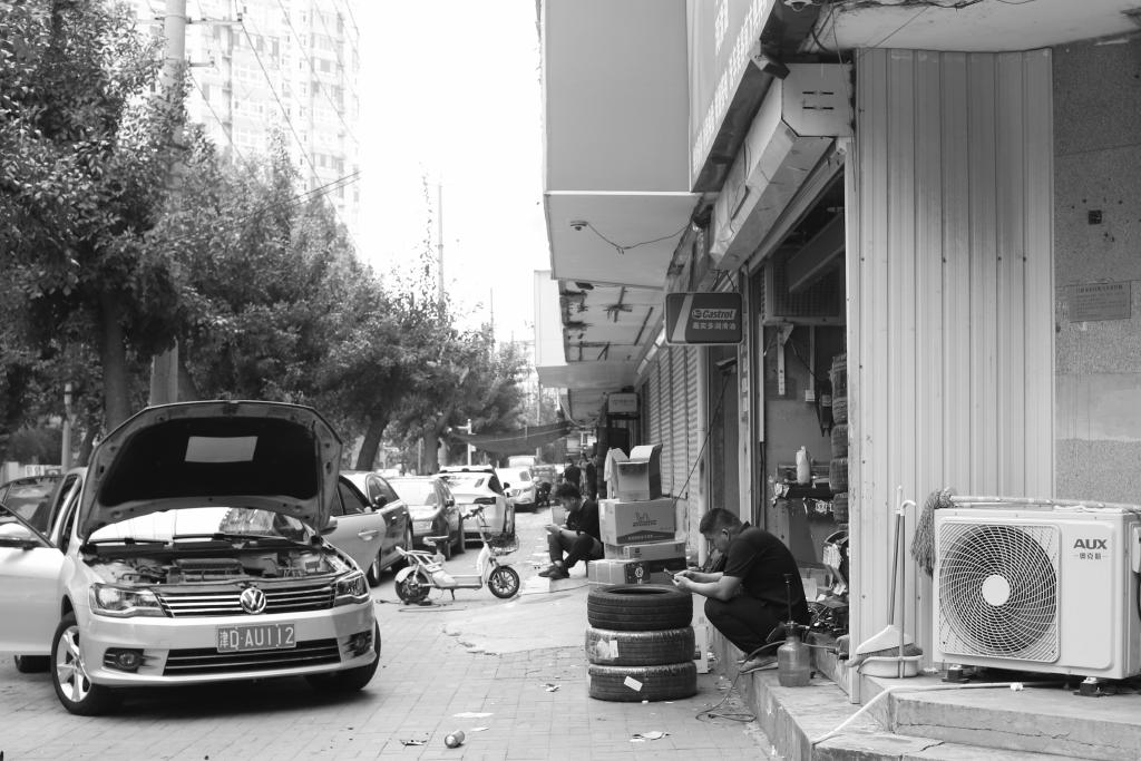 A black and white photo of a street in Tianjing (天京), China. On the left of the image a car sits parked, the passenger side door open and the bonnet (hood) propped up. On the right, a mechanic sits by a pile of three tyres on the steps leading to his workshop. His focus is on the phone in his hands. In the distance, a man sits on the steps of the next shop. He is also looking at his phone.