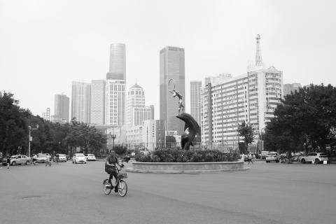 A black and white photo of a roundabout in Tianjing (天京), China. In the foreground, a lone cyclist occupies the roundabout on a rental bike. A statue stands in the middle of the roundabout.Tall skyscrapers line the horizon in the distance.