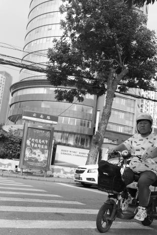 A black and white photo taken on the streets of Tianjing (天京), China. The photo is take from low down, looking up across a pedestrian crossing.  A man on an electric scooter looks purposfully at the camera as he enters the crossing. On the far side of the street, a curved glass building rises up to the top of the frame.