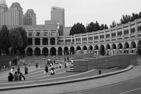 A black and white photo of Mingyuan Stadium (民园广场) in Tianjing (天京), China. The stadium has been turned into a public square and people can be seen sitting on steps that run round the inside of the stadium. Around the outer edge of what looks like a running track, an arched covering provides shade from the heat. The stadium feels like it was once a hive of activity, today it feels as if there is something missing.