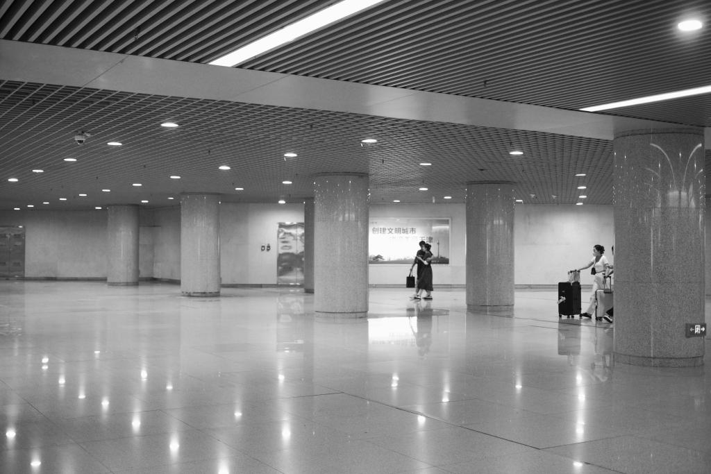 A black and white photo of the subway station in Tianjing (天京), China. The station is empty save for three people making their way across the hall with their lugguage. The bright ceiling lights reflect in the polished floor.