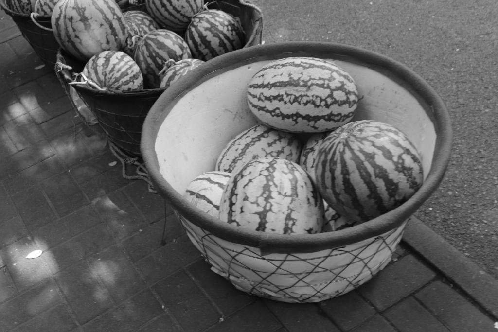 A black and white photo showing three baskets of watermellons lined up along the edge of a street in Tianjing (天京), China.