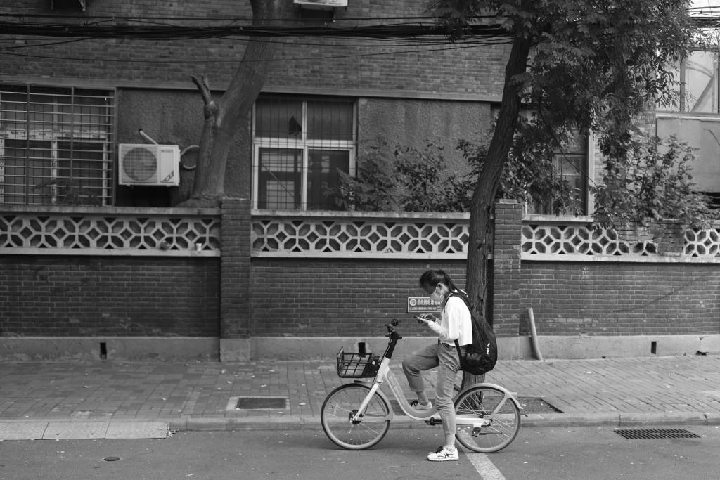 A black and white photo showing the profile view of a girl sitting on a rental bike on a street in Beijing. She has one foot on the pedal, the other rests on the ground steadying the bike. She glances down at her phone before setting off. Where to?