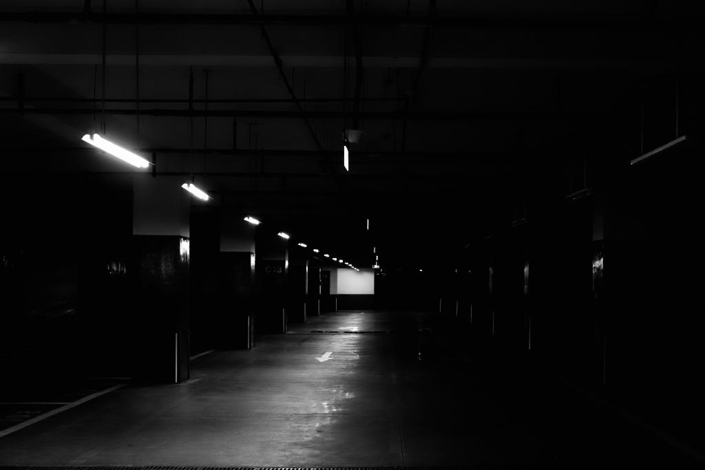 A black and white photo of an underground car park. The left half of the image is lit by artificial lights in the ceiling. The right half of the image is in darkness. No cars are parked in the parking bays. An arrow on the ground points towards the viewer.