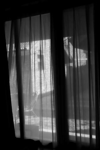 A black and white photo of our hotel courtyard taken through the bedroom window. Thin curtains hang infront of the window providing a translucent wave effect to the image. In the courtyard outside a chair can be seen. On the opposite courtyard the sunlight accents a brick wall.