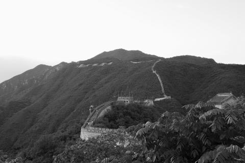 A black and white photo of the Great Wall of China. The wall starts in the lower centre of the frame and heads up the mountain side into the distance. There are two watchtowers or turrets visible in the photo. You can just make out a few people climbing the steeper sections of the wall.