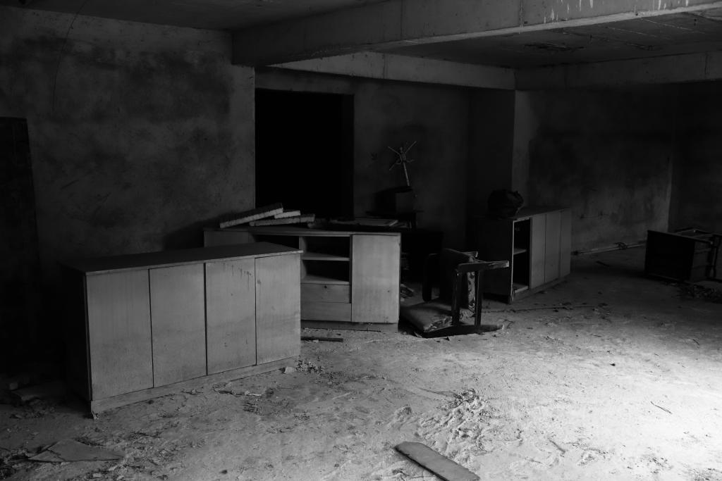 A black and white photo of an empty room. Discarded cupboards and an upturned chair litter the sandy floor. Light comes from behind the camera but isn't bright enough to penetrate a darkened doorway opposite. The bare walls show no indication as to whether this room has been lived in.
