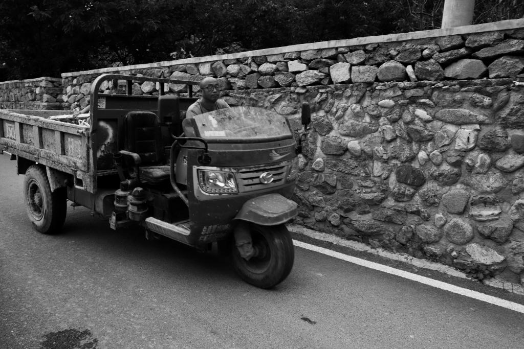 A black and white photo showing a man driving a three wheel flatbed truck. The vehicle has no cab and a half height windsheild. The man is looking straight ahead. Behind him a brick wall lines the road. Between the bars on the bed of the truck the face of a second person can be seen looking forwards along the road.