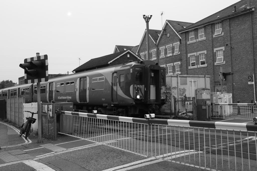 A black and white photo of a level crossing with barriers down across the road. A train coming from left to right across the image has just entered the crossing. On the far side of the tracks, brick building line the railway. On the near side, a bike rests against the warning lights.