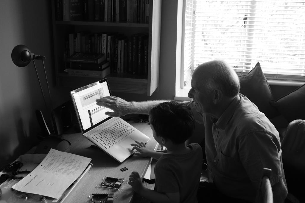 A black and white photo showing two people sat at a desk looking at a laptop screen. The older man points to something on the screen while a younger boy uses the trackpad. On the desk next to them are an SD card and two Raspberry Pi computers. The main light comes from a window behind them.