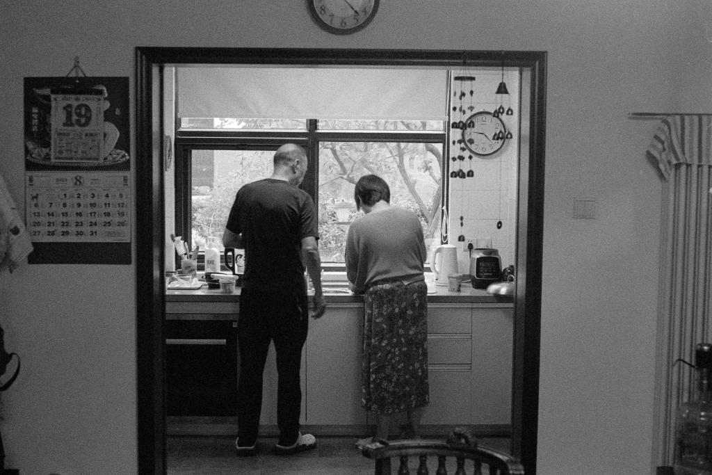 A black and white photo of the kitchen. This image was captured in Beijing in the height of summer. Two people stand next to each other looking at something on the counter in front of them. The clock on the wall shows that it is 9:24am. The image is a scan of a negative developed at home in Ilfosol3 (1+9).