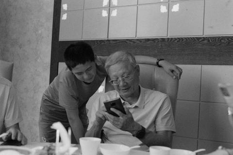 A grainy black and white photo of a grandfather and grandson sharing a moment together. The grandfather is seated and holding a phone. The grandson stands on his left looking over his shoulder at the phone, his arm around the chair. They are sat at a round dining table. Developed in Ilfosol 3 (1+9).